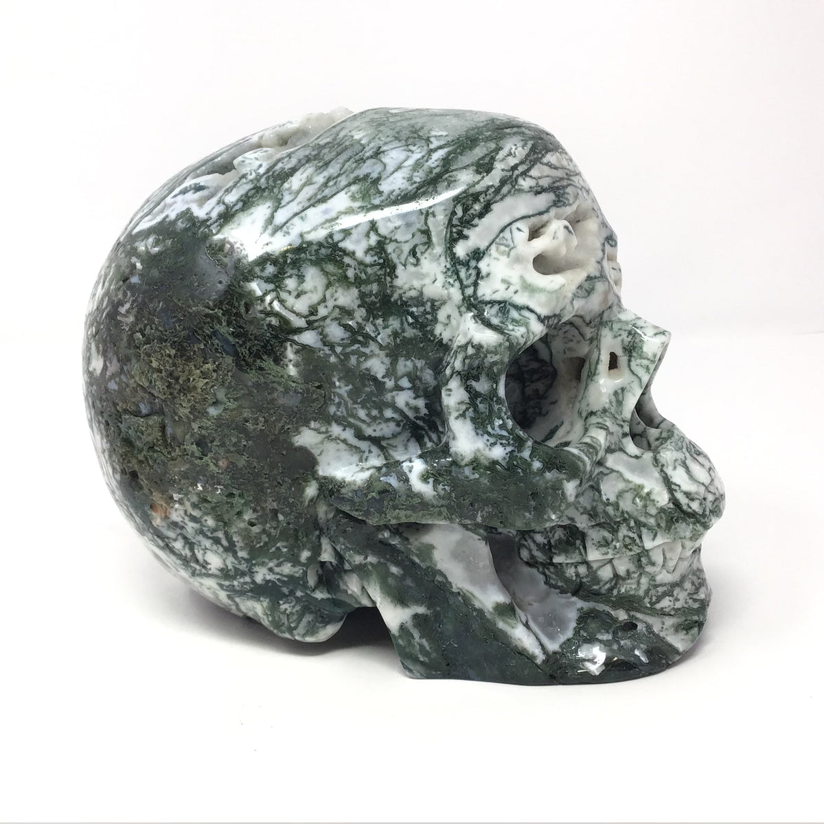 Green and white moss agate skull carving, angled side view showing mossy green veining and quartz druze