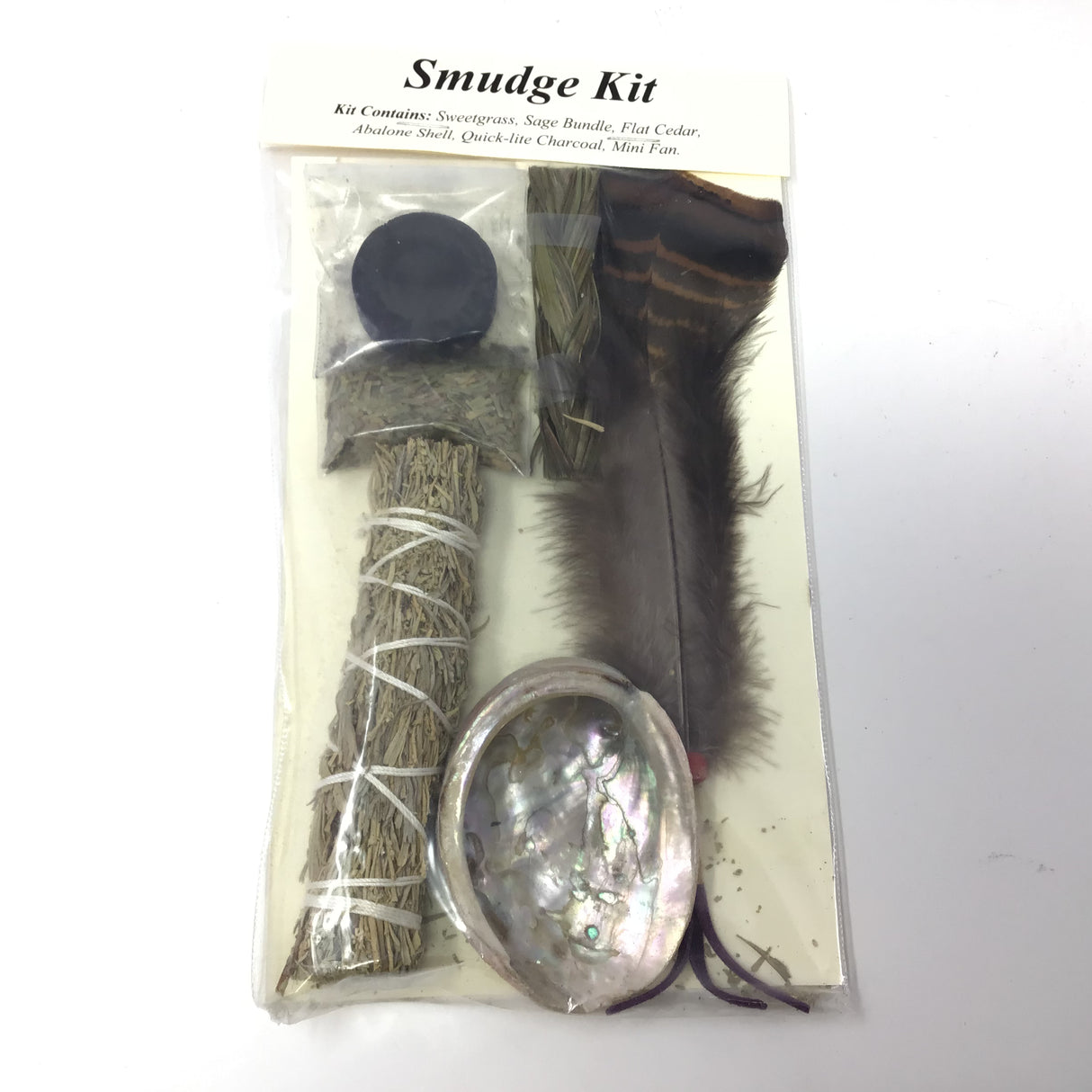 Smudge Kit contents through the clear pack: charcoal disc, sweetgrass braid, sage stick, shell and feather