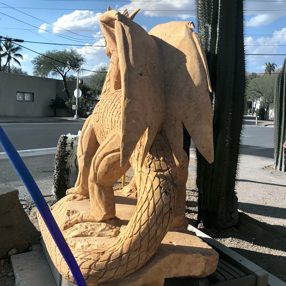 Hand-carved tan travertine dragon, large winged sculpture with scaled tail displayed outdoors under blue sky