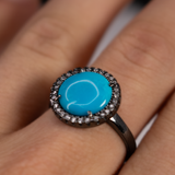 Sleeping Beauty turquoise cabochon ring with CZ halo and dark ruthenium band worn on a finger