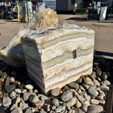 Zebra fossil onyx block fountain with 2 holes set in a river rock basin, horizontal cream and tan banding