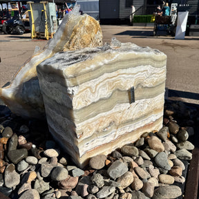 Zebra fossil onyx block fountain with 2 holes set in a river rock basin, horizontal cream and tan banding
