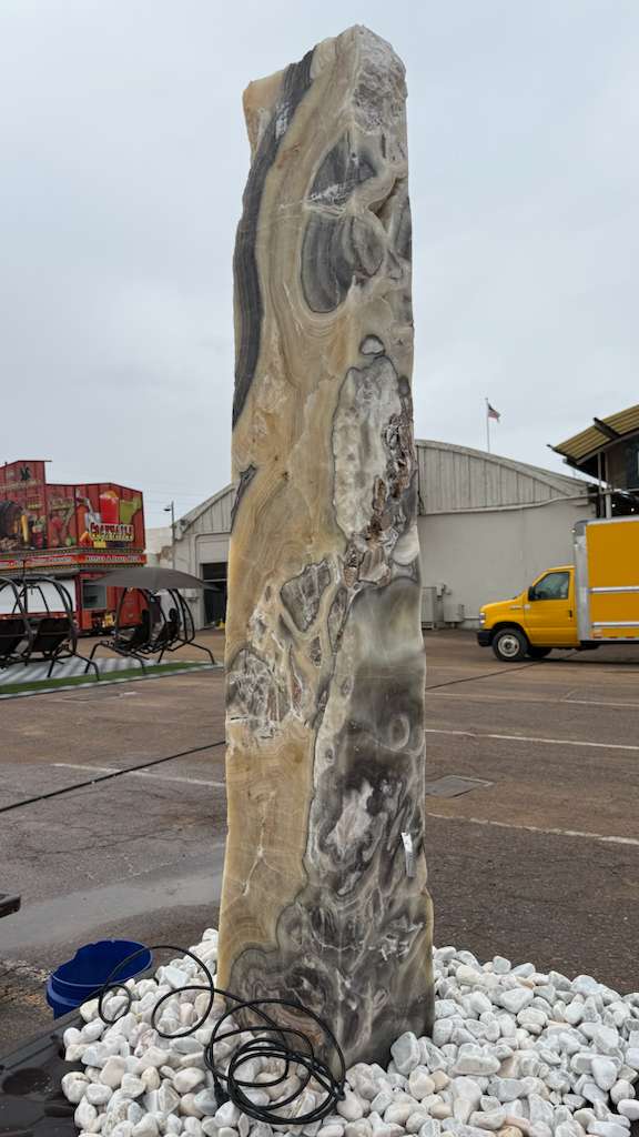 Full height cosmic onyx fountain pillar upright on white stones, cream and charcoal banding, tubing at its base