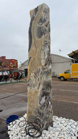 Full height cosmic onyx fountain pillar upright on white stones, cream and charcoal banding, tubing at its base