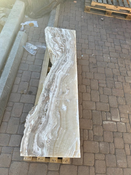 Vertical hourglass amber onyx fountain slab showing cream and tan banding, laid lengthwise on a pallet outdoors