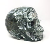 Green and white moss agate skull carving, angled side view showing mossy green veining and quartz druze