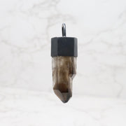 Smoky quartz crystal point pendant in oxidized black hexagonal cap bail on marble