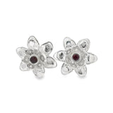 A pair of silver flower-shaped stud earrings with a small round dark red gemstone in the center of each flower, set against a plain white background.