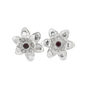 A pair of silver flower-shaped stud earrings with a small round dark red gemstone in the center of each flower, set against a plain white background.